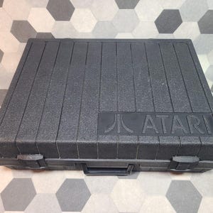 Può includere: Una valigetta rettangolare in plastica nera con superficie ruvida e scanalature verticali. La parola ATARI e il logo Atari sono impressi nell'angolo in basso a destra. La valigetta è dotata di una maniglia centrale e due chiusure a scatto.