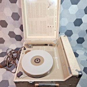 May include: A vintage Newcomb Solid State portable record player in a beige textured case. The unit features a white turntable platter, a brown power cord, and an open lid displaying printed operating instructions and the Newcomb Audio Products Co. logo.