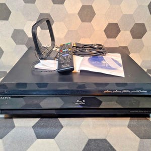 Pode incluir: Leitor Blu-ray Sony preto com controle remoto, antena e cabos. O leitor possui uma bandeja de disco e botões de função. O logotipo da Sony é visível na frente.