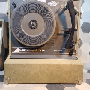 Op de afbeelding: Een olijfgroene vintage platenspeler met een donkergrijze draaitafel en een zwarte toonarm. Het label luidt "Audiotronics 300A". De unit heeft een getextureerde buitenkant en metalen hoekbeschermers. De draaitafel heeft snelheden van 16, 45 en 78 RPM.