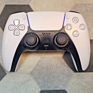 Puede incluir: Un mando de juego inalámbrico blanco y negro que cuenta con un panel táctil central, dos joysticks negros, una cruceta direccional y cuatro botones de acción marcados con símbolos de triángulo, círculo, cruz y cuadrado, con el logo PlayStation.