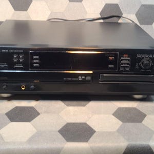 May include: A black Philips audio CD recorder with a silver logo. The device has a display screen, various buttons, and a disc tray. It features multiple input and output jacks and sits on four small feet. The background is a gray and white geometric pattern.