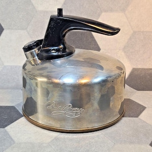 May include: A vintage silver-toned kettle with a black handle and spout. The kettle has a rounded body and a small spout with a lid. The handle is curved and the kettle has a base with a gold-colored rim. The kettle has the words "Paul Revere" engraved on the side.