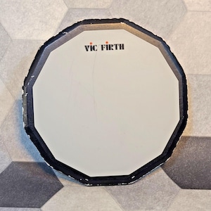 Könnte beinhalten: Ein weißes, achteckiges Vic Firth Übungspad mit schwarzem Rand. Das Vic Firth-Logo ist rot und schwarz aufgedruckt. Das Pad dient zum Üben des Schlagzeugspiels.