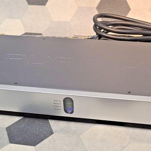 May include: A gray and silver Pure AV device with the brand name embossed on the top. The device has a silver front panel with indicator lights and a power button. A black cable is connected to the device.