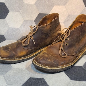 May include: A pair of brown leather ankle boots with light brown laces. The boots have a worn, distressed appearance with visible creasing and scuff marks. The soles are a darker brown with a cream-coloured trim.
