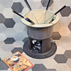 Könnte beinhalten: Ein dunkelgrauer Le Creuset-Fondue-Topf mit vier Gabeln mit schwarzen Griffen. Der Topf hat eine cremefarbene Innenseite und steht auf einem runden Sockel. Ein kleines Büchlein mit dem Titel "FONDUES" liegt neben dem Topf.
