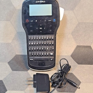 May include: A black Dymo label maker featuring a digital display screen, a QWERTY keyboard with number keys, and navigation buttons. The device includes a power button and various function icons. A black power adapter with a cord is included.