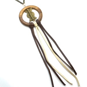 May include: A wooden pendant necklace with a circular wooden frame and a brass-colored decorative element in the center. Brown, cream, and brown leather strands hang from the bottom of the frame. The necklace chain is a dark bronze color.