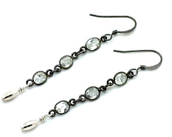 May include: A pair of dangle earrings with a dark metal chain and hook. Each earring features a series of oval-shaped, clear gemstones. The bottom of each earring has a silver-colored, elongated bead.