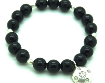 Può includere: Un braccialetto di perline nere con un ciondolo d'argento. Il braccialetto presenta grandi perline nere lucide infilate insieme con maglie d'argento. Un ciondolo rotondo d'argento con un motivo decorativo pende dal braccialetto.