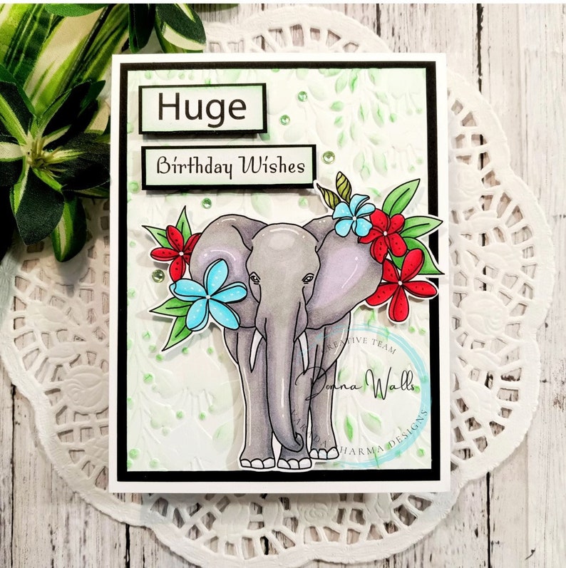 May include: A greeting card with a black border features a cartoon illustration of a gray elephant with pink ears and a floral arrangement of red, blue, and green flowers. The card has the text "Huge Birthday Wishes" in a black rectangle.