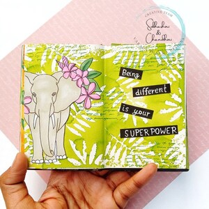May include: An open journal with a green background and white leaf patterns. The left page features a hand-drawn illustration of an elephant with pink flowers. The right page has the text "Being different is your SUPER POWER".