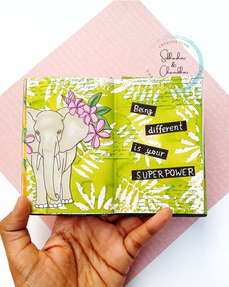 May include: An open journal with a green background and white leaf patterns. The left page features a hand-drawn illustration of an elephant with pink flowers. The right page has the text "Being different is your SUPER POWER".
