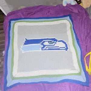 May include: A handmade crochet blanket featuring the Seattle Seahawks logo in shades of blue, green, and gray. The blanket has a square shape with a multi-colored border. The blanket is displayed on a purple surface.