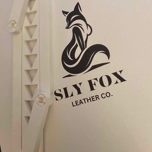 Puede incluir: Una superficie blanca con un logotipo de zorro estilizado negro y el texto "Sly Fox Leather Co."