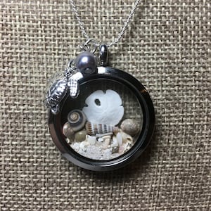 May include: A silver locket necklace with a glass charm containing sand, seashells, and a sand dollar. A silver turtle charm and a gray pearl dangle from the locket.