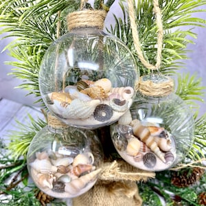 May include: Three clear glass Christmas baubles filled with sand and seashells. The baubles are hanging from a green artificial Christmas tree.
