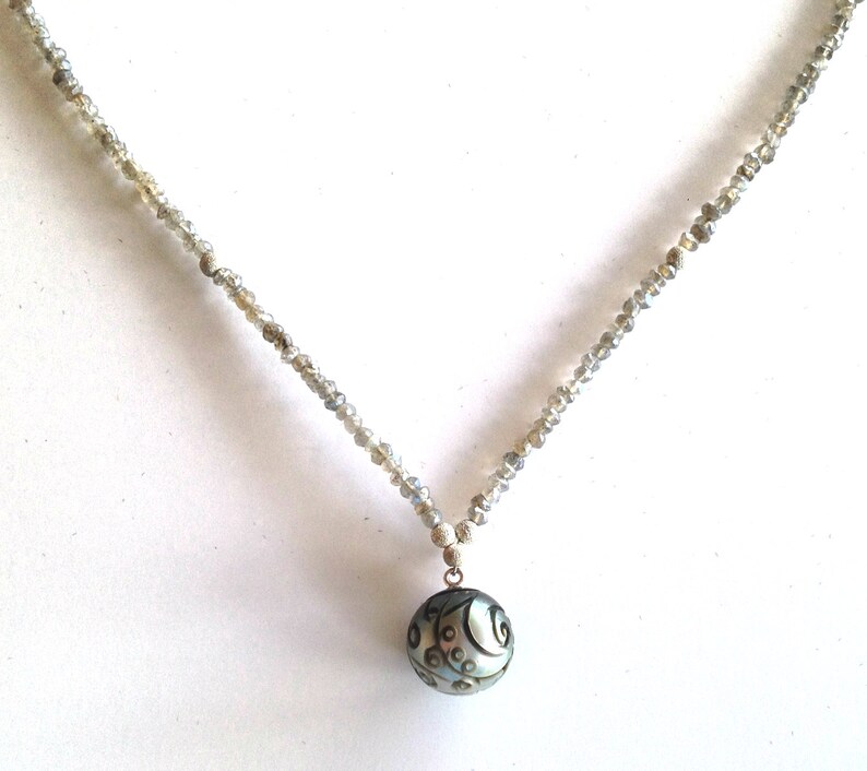 Puede incluir: Un collar de plata con un colgante redondo gris con un dise&ntilde;o de espiral. El collar est&aacute; hecho de peque&ntilde;as cuentas facetadas.