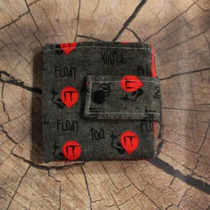 May include: A gray fabric wallet with a red and black design. The wallet features the words "FLOAT" and "YOU'LL FLOAT TOO" in white, along with red "IT" balloon graphics. The wallet has a snap closure.