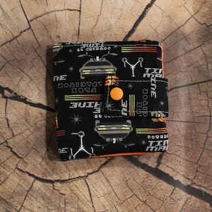May include: A black wallet with a repeating pattern of a DeLorean car, text, and other graphics in white, orange, and yellow. The wallet has an orange button closure and is set against a wood grain background.