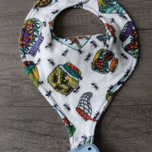May include: A white baby bib with a colorful print of jars and insects. The bib has a terry cloth border and a snap closure. A blue pacifier is attached to the bib.
