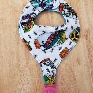 May include: A white baby bib with a blue trim and a pattern of insects in jars. The bib has a blue and white strap with a pink pacifier attached.