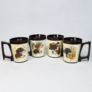 May include: A set of four ceramic mugs with dark brown rims and angular handles. Each mug features a cream-colored body decorated with detailed illustrations of various dog breeds, including pointers and retrievers, set against natural outdoor scenes.