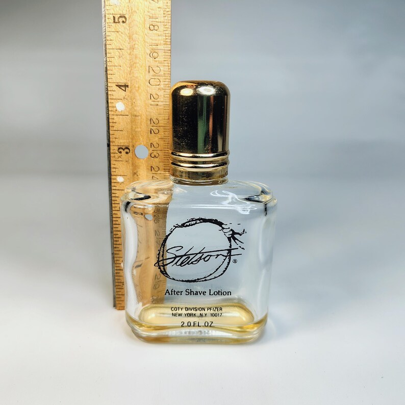Pu&ograve; includere: Una bottiglia di vetro trasparente di lozione dopobarba Stetson con tappo dorato. La bottiglia ha una forma rettangolare e contiene un liquido giallo. L'etichetta recita "Stetson After Shave Lotion" e "2.0 FL. OZ."