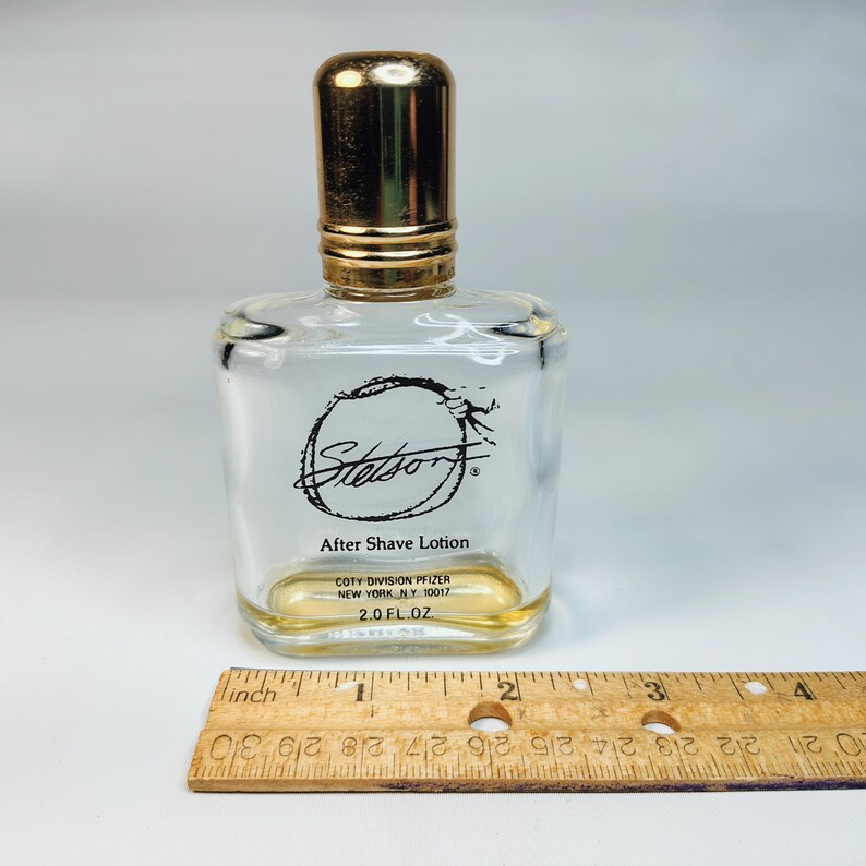 Pu&ograve; includere: Flacone di dopobarba Stetson in vetro trasparente con tappo dorato. Il flacone ha una forma rettangolare con bordi arrotondati e un logo nero. Contiene 59 ml di dopobarba. Un righello di legno &egrave; in primo piano.
