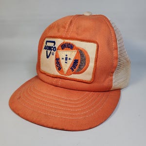 May include: Orange trucker hat with a white mesh back and a curved brim. The hat features a patch with the text "ARMCO V" and "UNION ROPE WIRE" in blue and orange. A vintage-style cap.