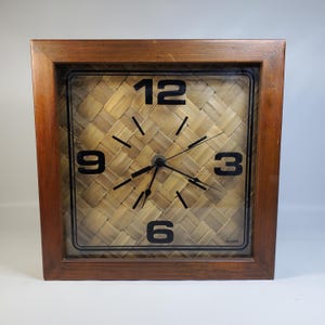 Puede incluir: Reloj cuadrado de madera con una esfera tejida de color marrón claro. Los números negros marcan las horas 3, 6, 9 y 12. Las manecillas del reloj son negras. El reloj está en un marco cuadrado de madera marrón oscuro. La marca "Arcante" está impresa en la parte inferior.