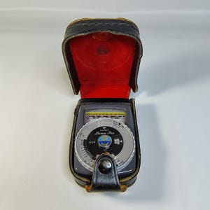 May include: A vintage Gossen Luna-Pro light meter in a black leather case with a red velvet interior. The meter has a silver dial with black and white markings and the text "Luna-Pro". The case is open, revealing the meter's face.