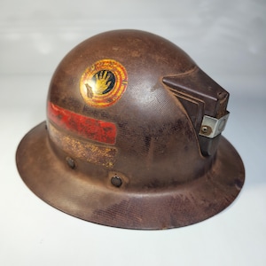 Può includere: Un casco di sicurezza marrone vintage con tesa larga e parte superiore arrotondata. Presenta un adesivo rosso e oro con un simbolo della mano e una striscia rossa. Il casco ha una visiera integrata e una clip metallica.