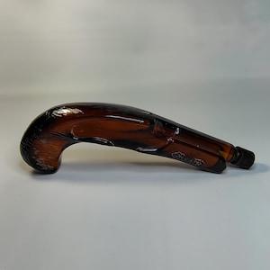 May include: A brown glass bottle shaped like a pistol. The bottle has a textured handle and a smooth barrel. The bottle has a black cap and is likely a decorative item or a container for liquids.