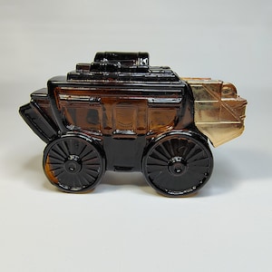 May include: A decorative stagecoach-shaped bottle. The bottle is primarily brown with black wheels and accents. The rear of the stagecoach is gold-colored. The design includes detailed wheels and a roof, creating a vintage aesthetic.