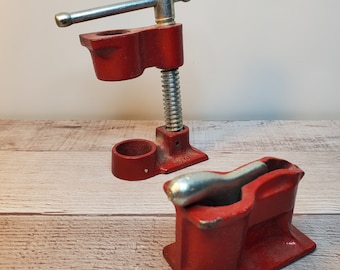 May include: A red metal clamp with a silver handle and a threaded screw. The clamp is designed to hold two pieces of wood together.