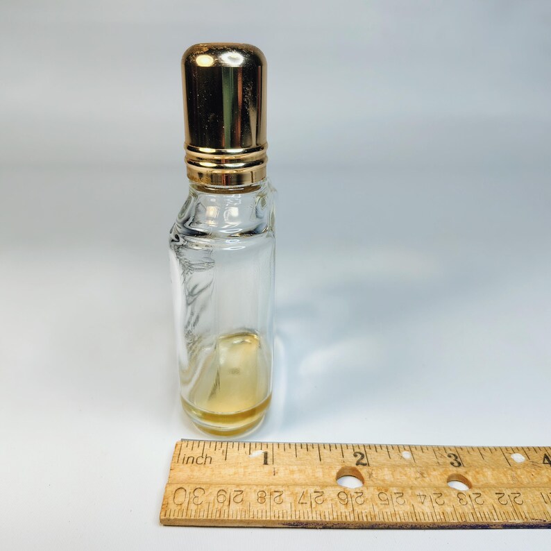 Pu&ograve; includere: Un piccolo flacone di profumo in vetro trasparente con tappo e dettagli dorati. Il flacone contiene una piccola quantit&agrave; di liquido color ambra. Un righello di legno &egrave; accanto al flacone.