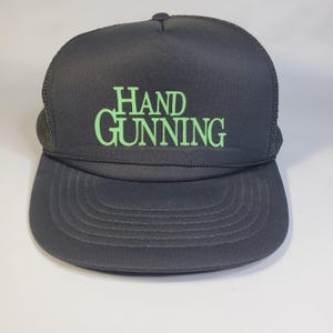 以下が含まれることがあります： 黒いトラッカーハット。つばはカーブしており、背面はメッシュ素材。前面には「HAND GUNNING」の文字が明るい緑色のブロック体でプリントされています。サイズ調整可能なスナップ留め付き。