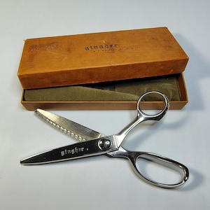 May include: Open vintage Gingher scissors with a serrated blade, displayed in an open brown box with the text "gingher, a tradition of quality". The scissors are silver-toned and rest on a green velvet interior.
