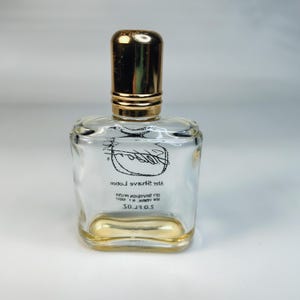 Pu&ograve; includere: Una bottiglia di vetro trasparente con un tappo dorato e una base dorata. La bottiglia ha una forma quadrata con bordi arrotondati. L'etichetta recita "After Shave Lotion" e "S.O.S."