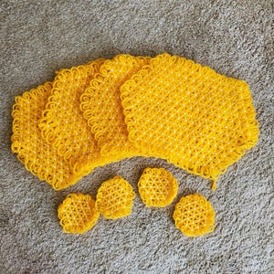 May include: A set of handmade yellow crochet kitchen items, including four square hot pads and four round coasters. The crochet work features a floral pattern with white accents. The items are arranged on a neutral-colored surface.