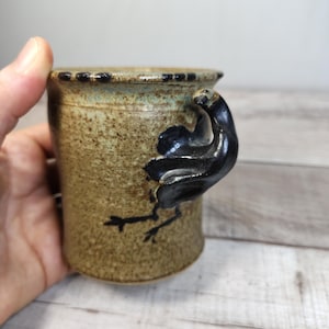 May include: A speckled tan ceramic mug featuring a dark brown, sculpted bird-shaped handle. The rim of the mug is accented with a series of small, dark brown dots, and the body has a textured, earthy finish with a glossy glaze.