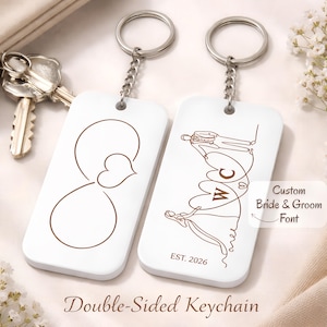 Puede incluir: Dos llaveros blancos de doble cara con anillas plateadas. Uno presenta un símbolo de infinito con un corazón, el otro tiene un diseño de novios con las iniciales "W C" y el año "2026".