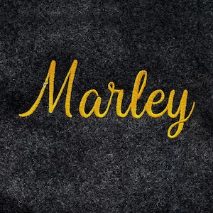 Könnte beinhalten: Nahaufnahme eines dunkelgrauen Stoffes mit dem Namen "Marley" in leuchtend gelber Schreibschrift gestickt. Der glänzende Stickfaden hebt sich vom strukturierten Hintergrund ab.
