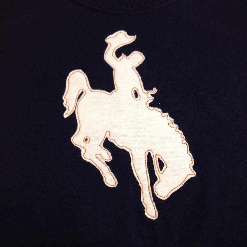 以下が含まれることがあります： 濃い青色のTシャツに、白い刺繍で描かれた、跳ね上がる野生の馬のモチーフ