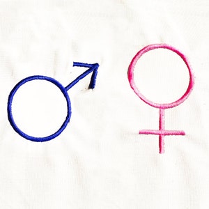 Puede incluir: Símbolos de género bordados sobre un fondo de tela blanco. El símbolo masculino es azul y el símbolo femenino es rosa.