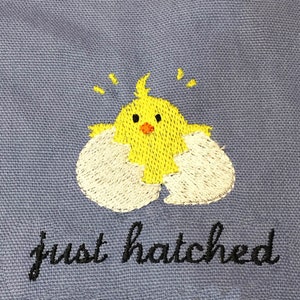 May include: Embroidered design of a yellow chick hatching from a white eggshell on a blue fabric background. The text "just hatched" is written in black cursive below the chick.