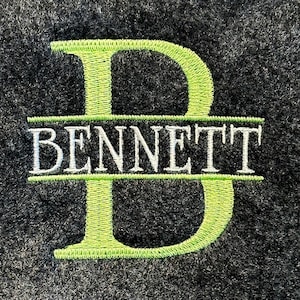 May include: Close-up of a dark gray, textured surface with the name "BENNETT" embroidered in white, framed by a large, lime green letter "D". The embroidery has a detailed, stitched appearance.