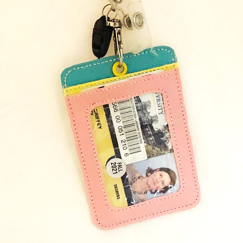 May include: A pink leather ID card holder with a clear plastic window. The holder has a yellow, blue, and green trim. The ID card inside the holder has a barcode, the text "WESTERN LEON LIBRARY SYSTEM", and the number "566 00 051 210 6". The ID card also has a photo of a person and the text "FALL 2021" and "39348093".